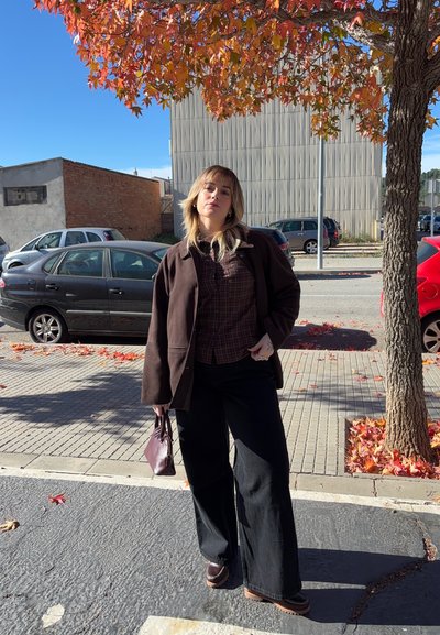 Chaqueta marrón de gran tamaño, camisa de cuadros, pantalones negros anchos y zapatos marrones. Sosteniendo un bolso pequeño color burdeos. Hojas otoñales y cielo despejado de fondo.