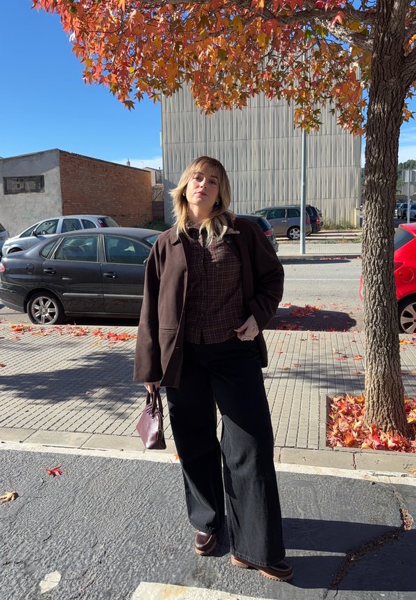 Bruine oversized jas, geruit overhemd, wijde zwarte broek en bruine schoenen. Houdt een kleine bordeauxrode tas vast. Herfstbladeren en een heldere lucht op de achtergrond.