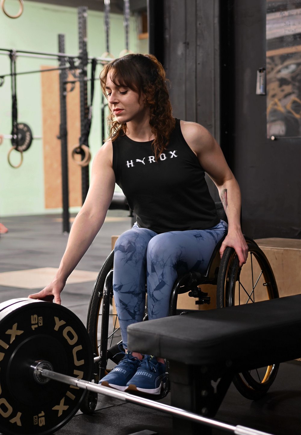Utilisateur de fauteuil roulant dans une salle de sport, portant un haut sans manches noir et un legging à motifs bleus, tendant la main vers une plaque de poids.