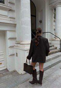 Blazer oversized marrón oscuro con hombros estructurados, combinado con botas de punta marrones hasta la rodilla y un bolso a juego, de pie en escalones de piedra.