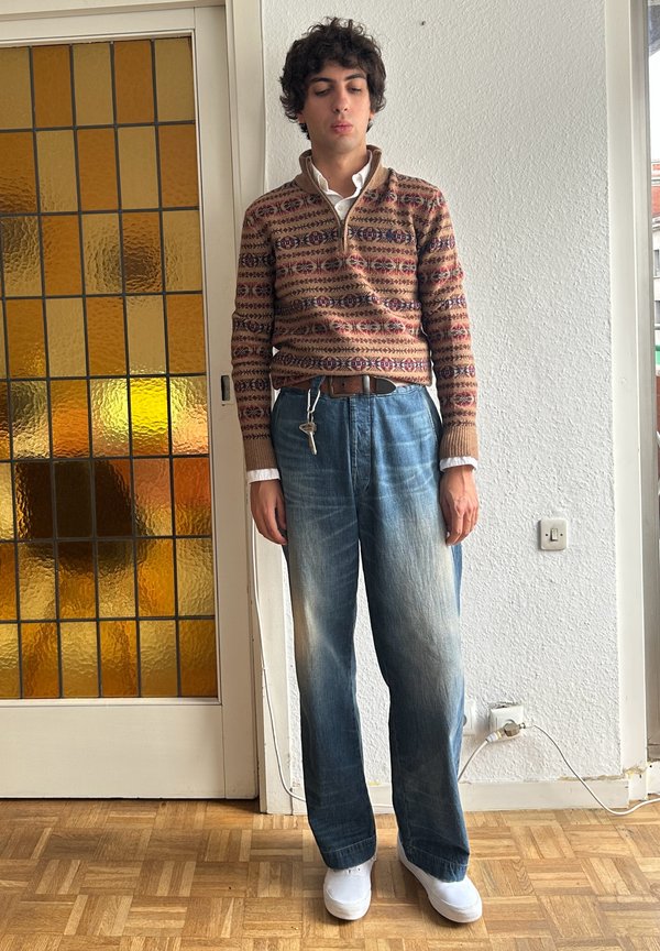Persona joven con cabello rizado que lleva un suéter marrón con estampado, camisa blanca, vaqueros anchos azules, zapatos blancos, sosteniendo llaves, de pie dentro de una habitación junto a la puerta.
