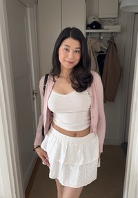Weißer, schichtweise aufgebauschter Rock mit floralem Muster und Rüschensaum, kombiniert mit einem weißen Tanktop und einer sanft rosa Strickjacke, dazu eine schwarze Umhängetasche.