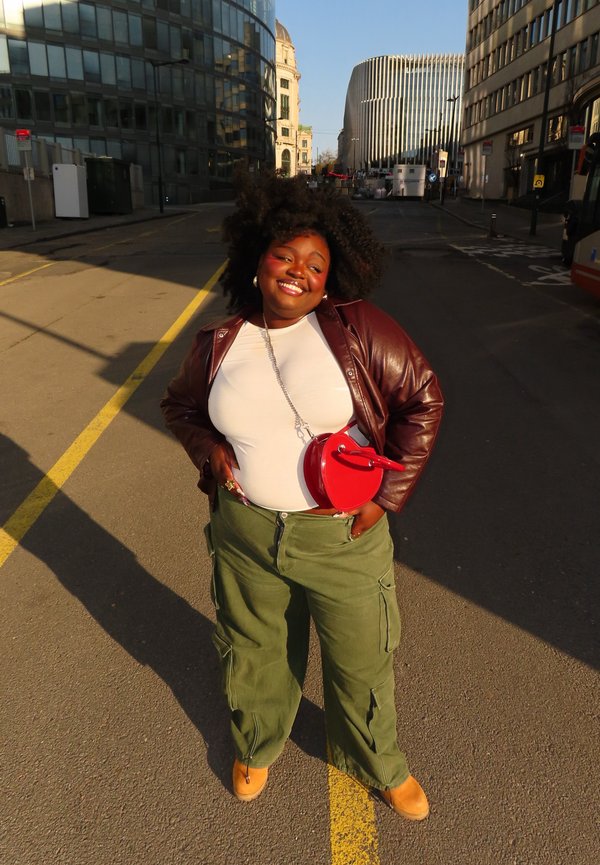 Mosolygó, göndör hajú nő áll egy városi utcán, fehér felsőt, bordó dzsekit, zöld terepmintás nadrágot, világosbarna csizmát és piros szív alakú táskát viselve.