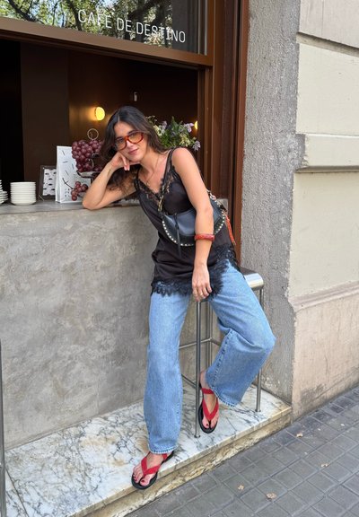 Camiseta de tirantes con encaje negro, jeans anchos de color azul claro, chanclas rojas. Encimera de mármol, fondo de café de madera con uvas y flores.