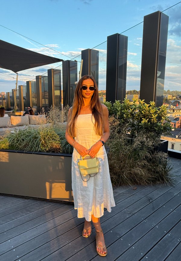 Una mujer con gafas de sol y un vestido blanco sostiene un bolso pequeño en una terraza en la azotea con jardineras al atardecer.