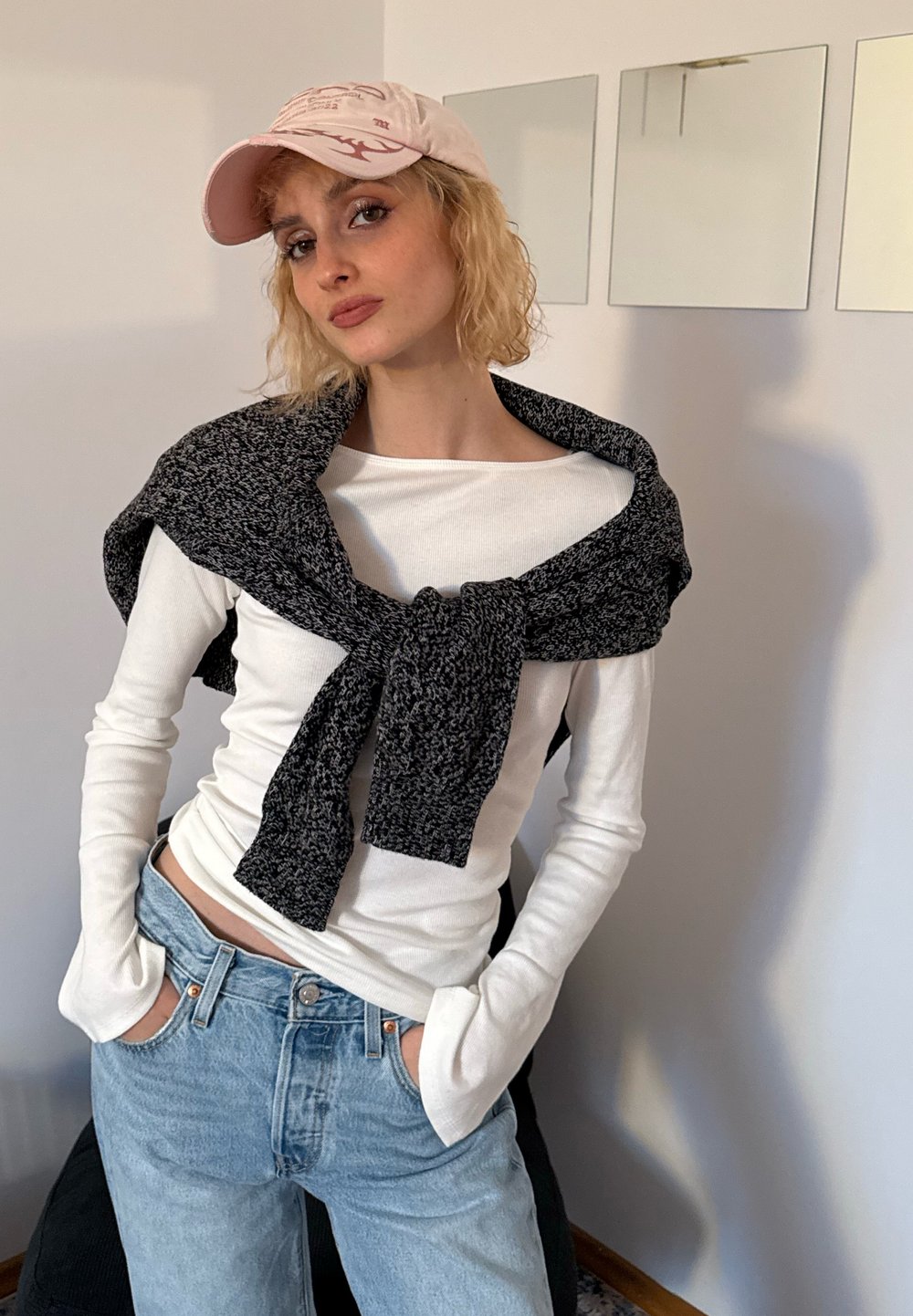 Camicia bianca a maniche lunghe con un maglione lavorato a maglia nero e grigio annodato attorno al collo, abbinata a jeans a vita alta di colore azzurro chiaro. Tessuti morbidi e stile casual.