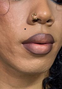 Nærbilde av lepper med en gradient av malva- og taupe-nyanser, supplert med et lite svart skjønnhetsmerke og en gyllen septumring.