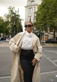Trench coat bej peste o cămașă albă cu guler ondulat, asortat cu o fustă neagră cu dungi verticale. Accesorii includ ochelari de soare negri supradimensionați și o poșetă bej.