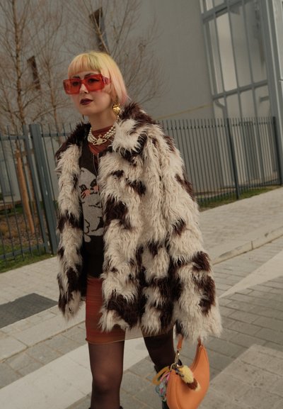 Persona con cabello rubio y gafas de sol naranjas que lleva un abrigo esponjoso con estampado, suéter gráfico y sostiene un bolso naranja al aire libre.