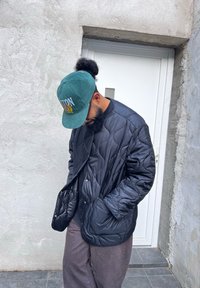 Μαύρο ραμμένο μπουφάν με γυαλιστερή επιφάνεια, με στρογγυλή λαιμόκοψη και μπροστινές τσέπες, φορτωμένο πάνω από σκούρες γκρι παντελόνες και ένα πράσινο καπέλο.