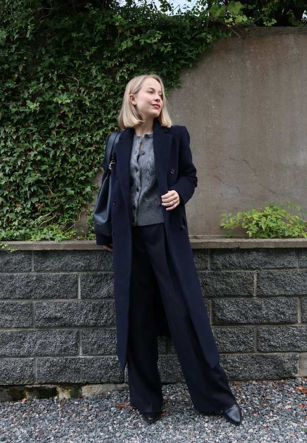 Una mujer rubia con un abrigo azul marino y pantalones de pernera ancha posa junto a una pared cubierta de hiedra y lleva un bolso negro al hombro.