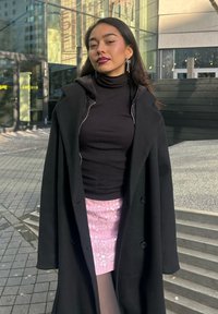 Schwarzer Wollmantel mit Kapuze, getragen über einem schwarzen gerippten Top. Pinker Pailletten-Minirock. Modell hat lange schwarze Haare und auffällige Ohrringe.