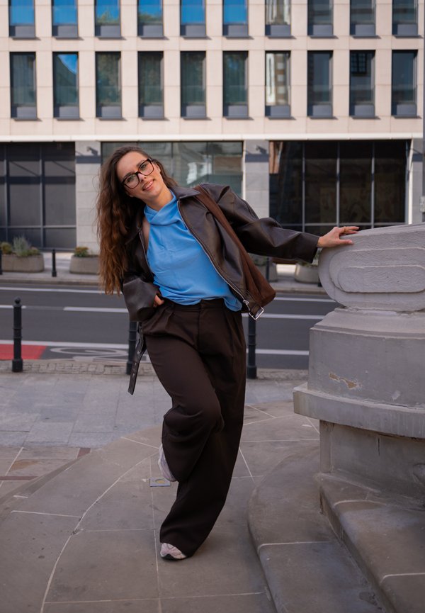 Joven mujer con cabello largo y gafas se apoya en una barandilla de piedra al aire libre, vistiendo una camisa azul, chaqueta marrón y pantalones negros.