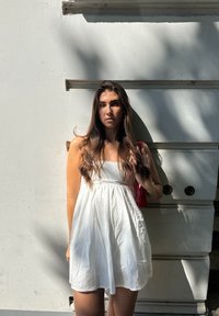 Robe blanche sans manches avec une taille froncée et une jupe fluide ; le mannequin tient un petit sac rouge ; fond uni avec de légères ombres.