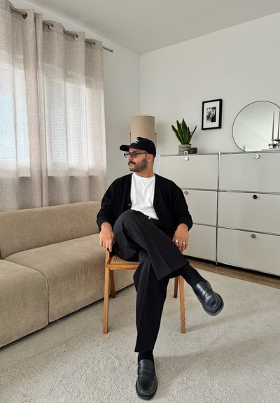 Hombre con gorra negra, gafas, camisa blanca y pantalones negros sentado con las piernas cruzadas en una silla de madera en una sala moderna con sofá beige.