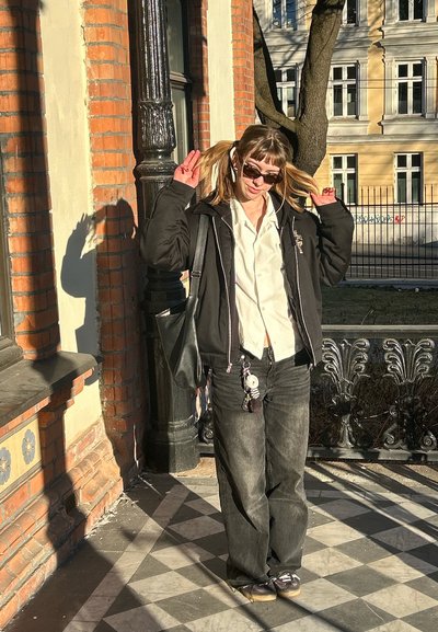 Chaqueta negra sobre una camisa blanca, jeans negros y zapatillas. Cabello ondulado suelto con gafas de sol. Sombra proyectada sobre un suelo texturizado.