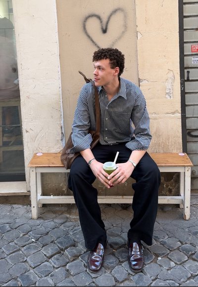 Camisa de cuadros azul y blanca, pantalones oscuros, bolsa de mensajero marrón, sosteniendo una bebida verde en un vaso desechable, sentado en un banco de madera.