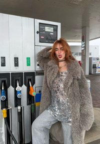 Manteau gris et duveteux porté sur un haut argenté orné de sequins, assorti à un jean gris clair. Plusieurs pompes à carburant avec des buses colorées en arrière-plan.
