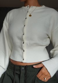 Weißer, gerippter Pullover mit rundem Ausschnitt und schwarzen Knöpfen, kombiniert mit grünen Hosen mit Kordelzug. Der Stoff wirkt weich und strukturiert.