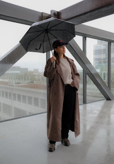 Abrigo de lana marrón con un corte holgado, suéter gris, jeans negros y botas. Sosteniendo un paraguas negro en interiores con un fondo de cristal.