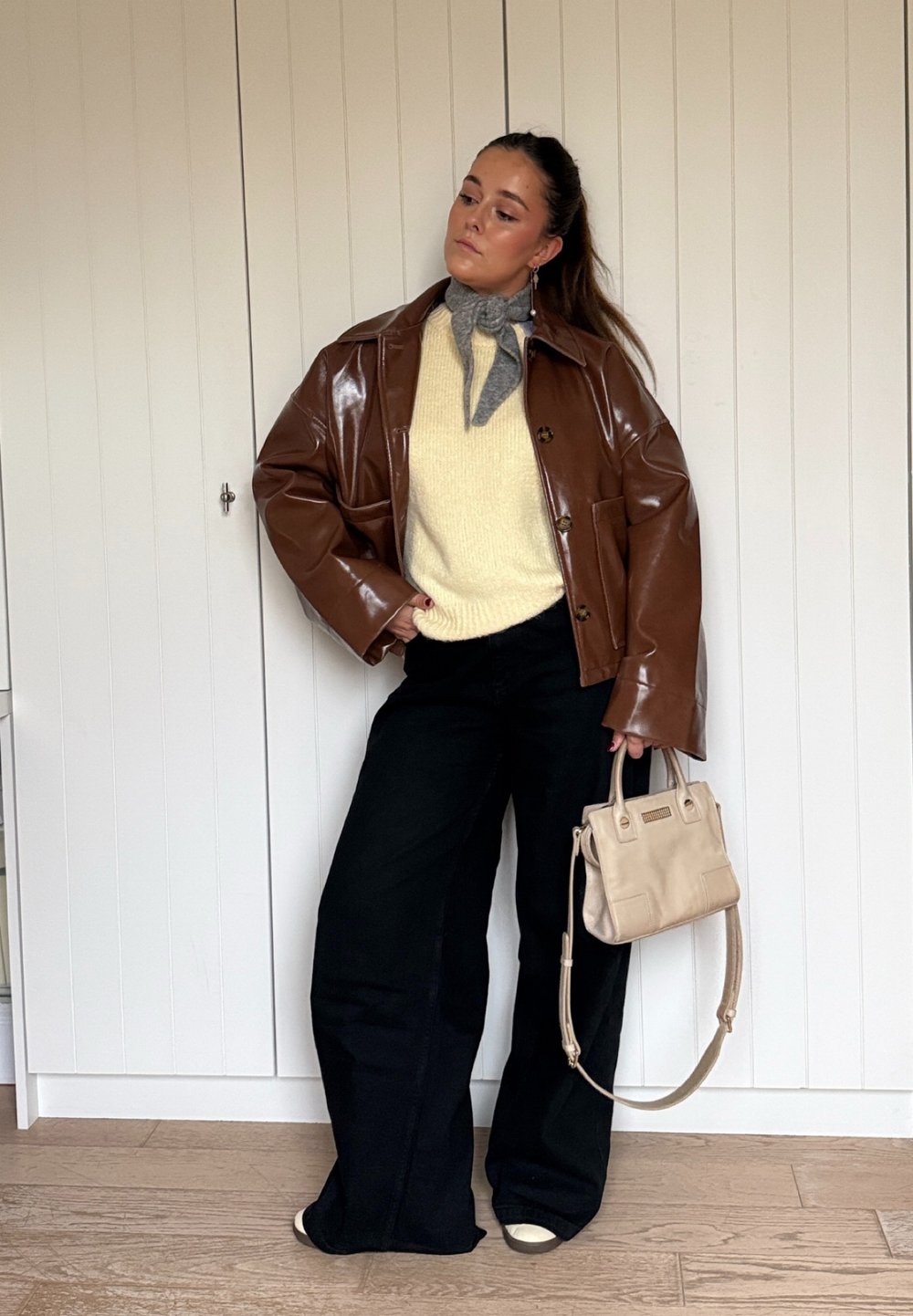 Brown glossy oversized jacket, cream knitted jumper, wide-leg black trousers, grey neck scarf, and a small beige handbag with short handles.