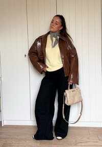 Brown glossy oversized jacket, cream knitted jumper, wide-leg black trousers, grey neck scarf, and a small beige handbag with short handles.
