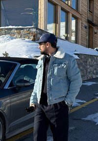 Veste en denim bleu clair avec un col en fausse fourrure blanche, portée sur une chemise noire. Il tient des gants noirs et se tient à côté d'une voiture grise.