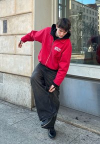 Κόκκινο zip-up μπουφάν με λογότυπο "STWD", μαύρη φόρμα από μέσα, φαρδιά σκούρα τζιν και μαύρα παπούτσια, ποζάροντας μπροστά από έναν πέτρινο τοίχο.