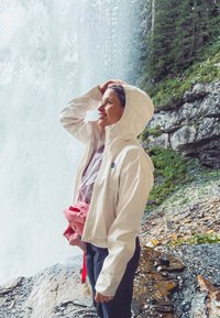 Hvit vanntett jakke med hette, lagdelt over en lett skjorte. Rosa midjebelte med stropp. Naturlig bakgrunn med et fossefall og steiner.