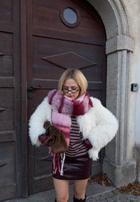 Cremefarvet jakke af imiteret pels, top med vinrøde striber, nederdel i bordeauxl�æder, stor ternet tørklæde i pink og bordeaux, brun håndtaske, mørke knæhøje støvler.