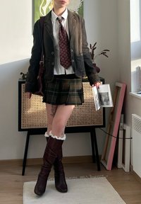 Brun kordfløyelsblazer, hvit skjorte, rødt mønstret slips, rutete miniskjørt, brun knelang støvler med hvite volanger, holder en bok.