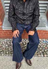 Čierna košeľa na gombíky so striebornými vertikálnymi pruhmi, v kombinácii s tmavomodrými rozšírenými džínsami a hnedými koženými čižmami, sediaci na dlažobných kameňoch.