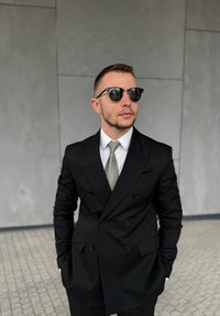 Mand i sort dobbeltradet jakkesæt med mønstret slips og solbriller, stående med hænderne i lommerne mod en grå flisevæg.