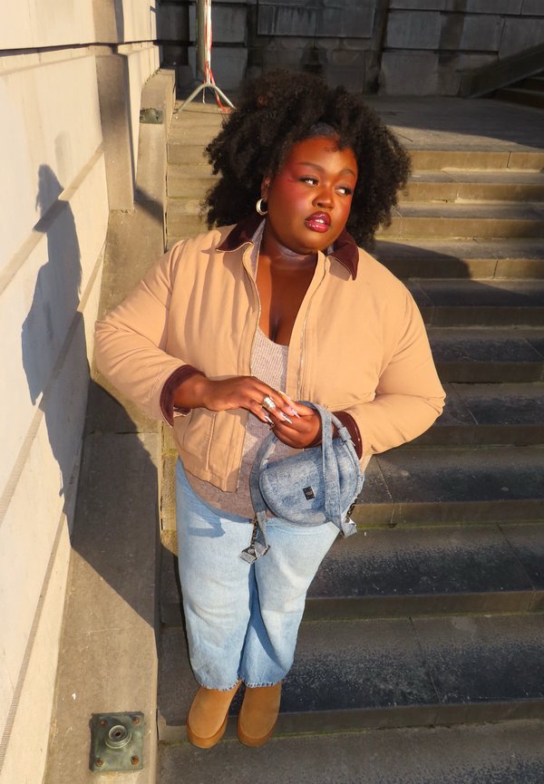 Göndör hajú nő áll kőlépcsőn, bézs dzsekit, világoskék farmert és barna csizmát visel, miközben egy kis farmer hátizsákot tart a napfényben.