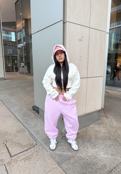 Sudadera corta blanca, pantalones de chándal rosas holgados y una gorra rosa. Zapatillas con acentos en gris claro y oscuro. Entorno urbano con baldosas de piedra.