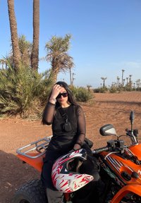 Un vivace quad arancione con un casco nero e rosso nelle vicinanze. L'utente indossa un top nero trasparente e pantaloni neri, seduto su un terreno sabbioso tra palme.