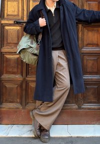 Personne portant un manteau bleu marine, un pull marron, un pantalon beige et des bottes marron, appuyée contre une porte en bois ornée, portant un sac à bandoulière vert.