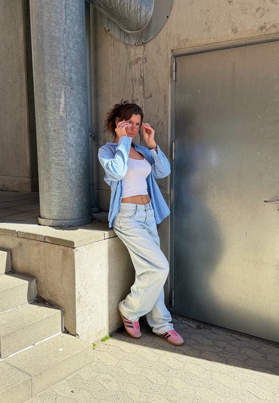 Mujer con camiseta blanca sin mangas, camisa azul a rayas, vaqueros claros y zapatillas rosas ajusta sus gafas de sol mientras se apoya en un saliente de hormigón cerca de una puerta metálica.