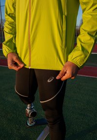 Chaqueta ligera de color lima con puños elásticos y cremallera. Mallas negras con detalles reflectantes y logo, mostrando una pierna protésica.