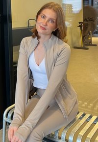Tenue de sport beige avec une veste zippée sur un haut blanc, assise sur un banc métallique dans un espace intérieur moderne.