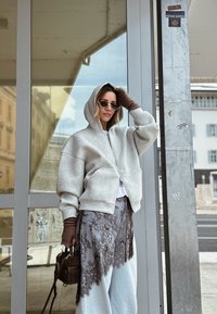 Femme en sweat à capuche gris ample, jupe avec superposition de dentelle, gants marron sans doigts, lunettes de soleil, tenant un sac à main marron, debout à côté de portes vitrées.