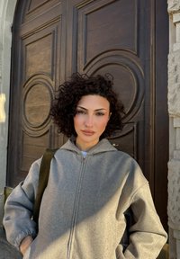 Kvinde med krøllet mørkt hår iført en grå hættetrøje med lynlås står foran en stor træd�ør med cirkulære udskæringer.