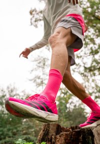 A person stepping outdoors onto a tree stump, wearing bright pink running shoes and matching socks, dressed in shorts and a long-sleeved top.