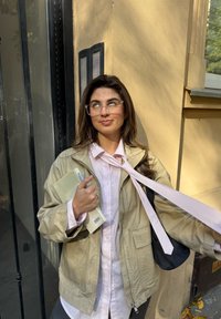 Smėlinė striukė virš šviesiai rožinės dryžuotos marškinės, laikantis užrašų knygutę ir rašiklį, su rožiniu kaklaraiščiu nuleistu ir juoda rankine ant peties.