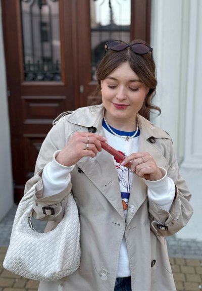 Gabardina beige, suéter blanco, tejido con estampado. Sosteniendo un producto de labios rojo con detalles plateados. Bolso de hombro de tejido blanco. Fondo de puerta de madera.
