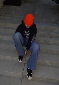 Melns kapuci džemperis ar grafisku attēlu, zilas plataji džinsi un melni sporta apavi ar baltām detaļām un brūnām zolēm. Spoži oranža cepure.