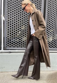 Hnedý oversize kabát s veľkými gombíkmi, biela košeľa, tmavohnedé kožené nohavice s rozšírením a špicaté lodičky na vysokých opätkoch. Textúrované pozadie.