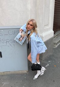 Lyshåret kvinde i blå skjorte og mininederdel holder et magasin og en sort håndtaske og har hvide sokker med sort-hvide prikkede hæle på gaden.