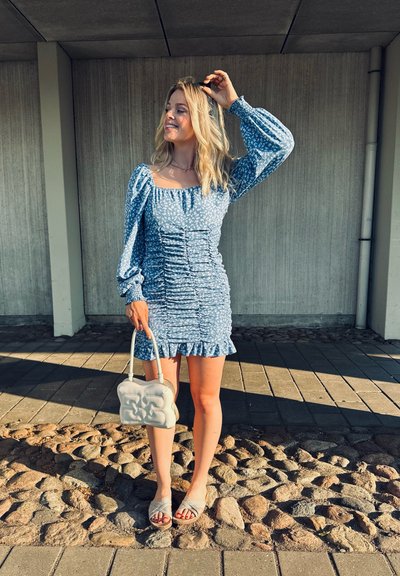 Vestido mini floral azul claro con detalles fruncidos, mangas largas y abullonadas, y dobladillo con volantes. Combinado con un bolso beige y sandalias planas.