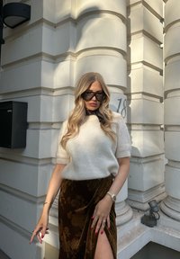 Bela pletena jopica in rjave sametne krilo z cvetličnim vzorcem. Sončna očala in črn šal kot poudarek. Kulisa vsebuje teksturirane bele stebre.